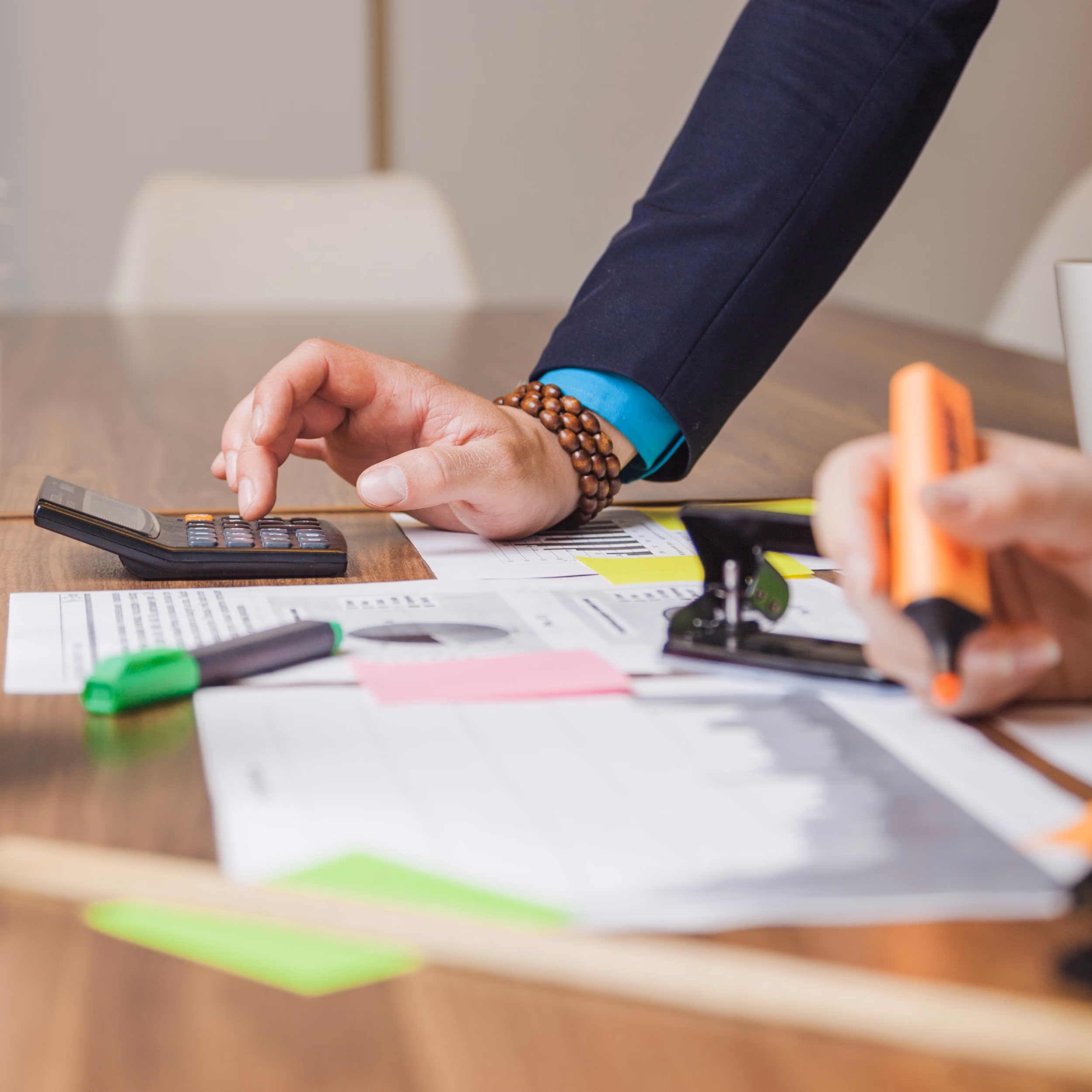 Person using a calculator whilst reviewing financial documents and papers with colourful sticky notes on a desk, with another person holding a highlighter in the background.