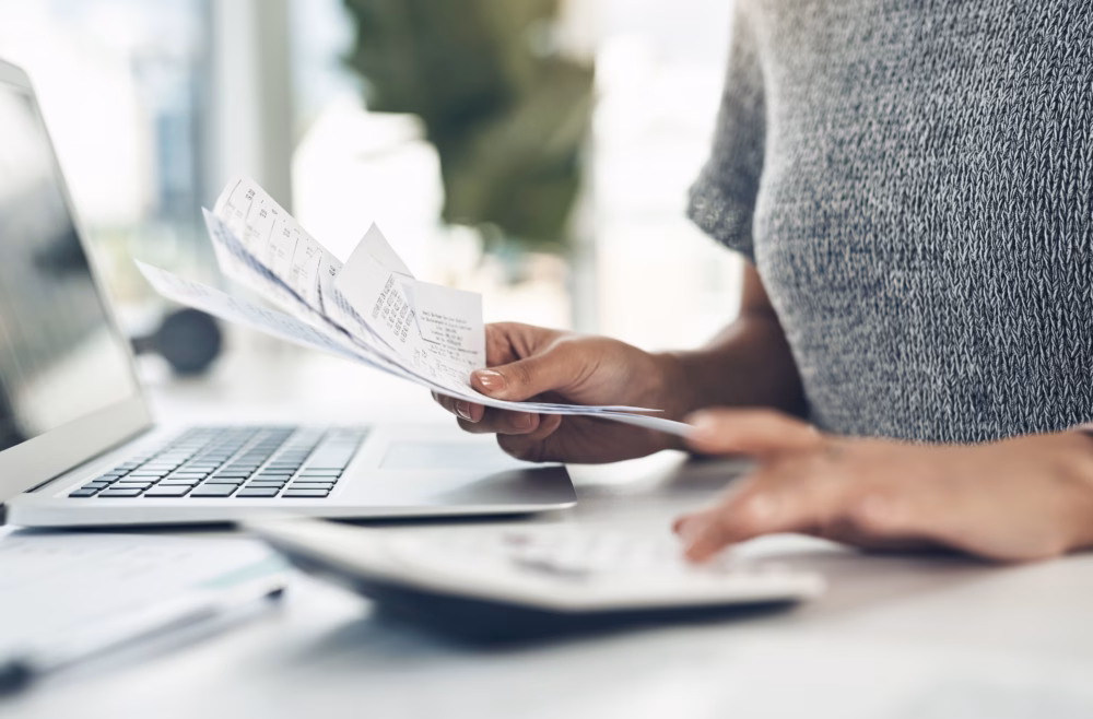Person in a grey jumper reviewing financial receipts or bills whilst working on a laptop, with paperwork on the desk.